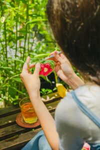 A woman enjoys crocheting outdoors in a lush garden, crafting a colorful piece with visible green leaves.
