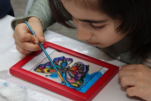A child focuses on painting a vibrant butterfly in a red frame indoors.