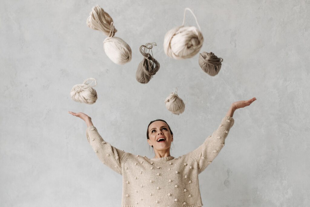 A woman in a cozy sweater joyfully tosses yarn bundles indoors, showcasing creativity and happiness.